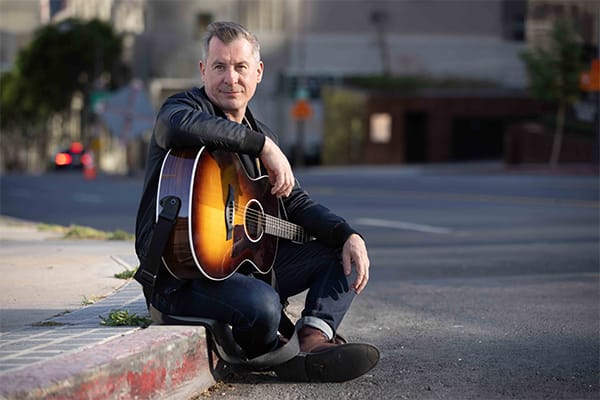 Bren Holmes sitting on curb with acoustic guitar